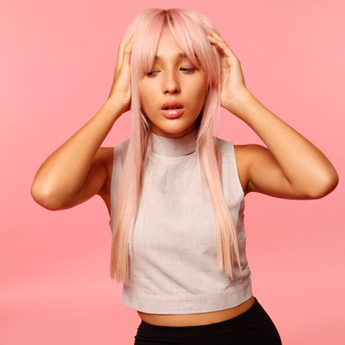 Girl with straight, pink hair and a pink T-shirt is standing with her hands touching her hair and looking away from the camera.