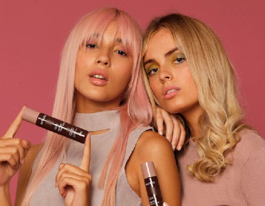 Girl with straight, pink hair is holding the Restora Protein Treatment between her two hands and standing beside a girl with curly, blonde hair who is resting her head on her shoulder and holding the Restora Protein Treatment in her right hand.